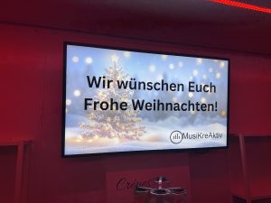 Schriftzug auf einem Bildschirm "Wir wünschen Euch Frohe Weihnachten" mit dem Logo der Musikschule.